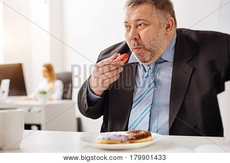 Too many sweets. Outsized compulsive unhealthy man sitting at his workplace and stuffing himself with baked sweets while looking rather unhealthy