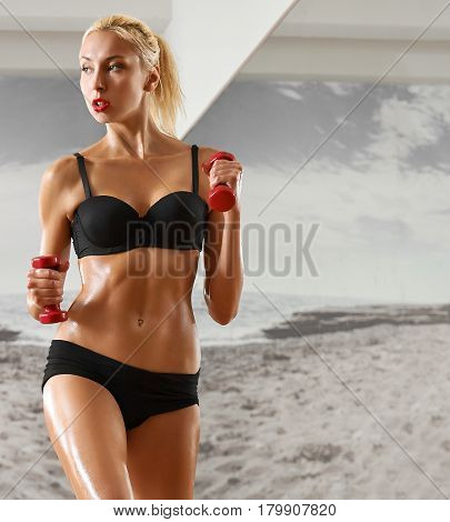 Sexy, athletic, blonde woman in the gym, against the background of the mirror, with dumbbells in hands. Sports, pumped press, a beautiful body, body Parts.