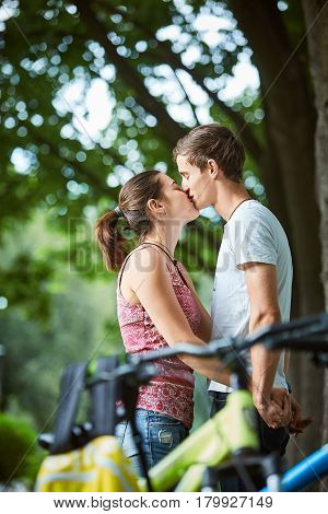 Young people, couple with bicycles in the park. A loving couple on a date on a summer evening. The guy with the girl hugs and kisses. Youth, first feelings, first love, first dates