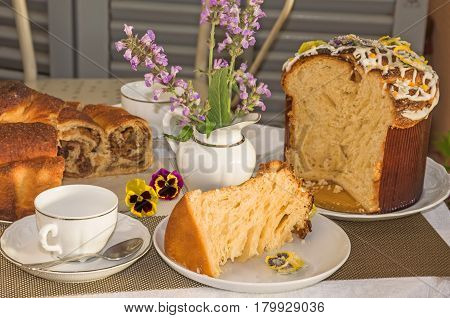 Typical italian flavorous home baked cakes for Easter: La Gubana with sophisticated stuffing and Panettone with candied flowers on the table covered for tea time. Horizontal close up. Main colors: white gray yellow brown.