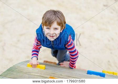 Happy blond kid boy having fun and climbing on outdoor playground. Funny joyful child smiling and romping on slide. Summer, spring and autumn leisure for active kids. Boy in colorful fashion clothes