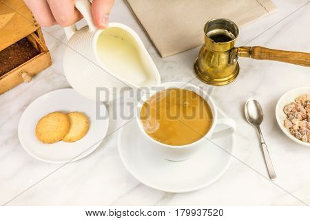 Milk just poured into a cup of coffee, with vintage coffee grinder and pot in the background, and cane sugar. Selective focus