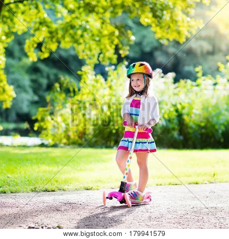 Little child learning to ride a scooter in a city park on sunny summer day. Cute preschooler girl in safety helmet riding a roller. Kids play outdoors. Active leisure and outdoor sport for children.