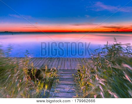 Long Exposure Image Of Blue And Orange Sunset Over Jetty On The Shore Of A Lake