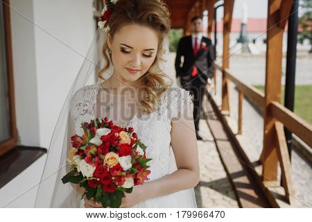 Gorgeous blonde bride posing with bouquet happy newlywed woman in white wedding dress holding roses bouquet groom looking in the background
