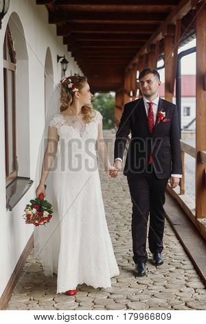 Happy bride holding hands with groom beautiful blone bride in white wedding dress walking with handsome groom on paving road near wall happy newlyweds