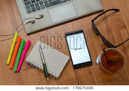 Nitra, Slovakia, april 3, 2017: Expedia application in a mobile phone screen. Workplace with a laptop, an earphones, notepad, pen, tea, sunglasses and color markers on wooden background