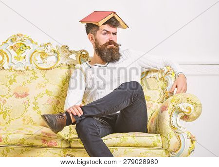 Macho Sits With Open Book On Head, Like Roof. Man With Beard And Mustache Sits On Sofa, White Wall B