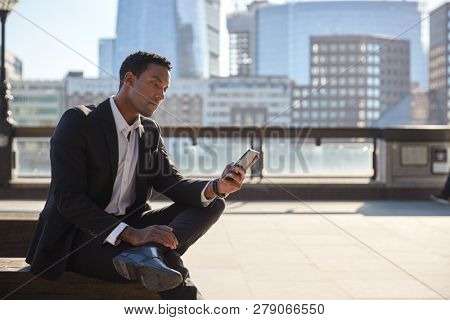 Millennial businessman wearing black suit and white shirt sitting on the River Thames embankment using smartphone, close up