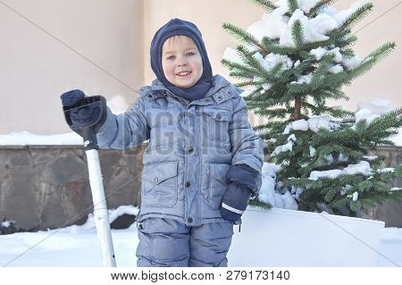 Cute Little Caucasian Baby Boy Shovels Snow In The Yard With Fir-tree On Background. Winter Outdoors