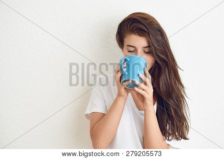 Young Brunette Woman In White T-shirt Drinking From Blue Mug With Both Hands, Looking At Camera With