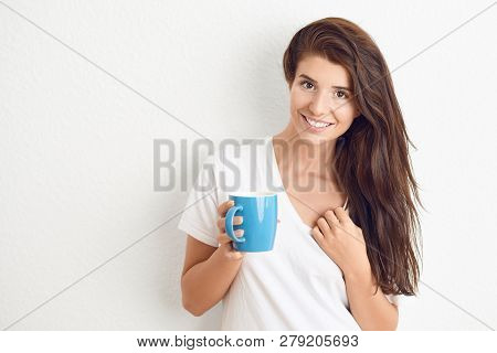 Young Brunette Woman In White T-shirt Drinking From Blue Mug With Both Hands, Looking At Camera With