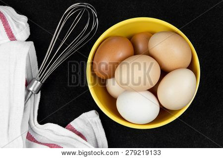 Top View Of Hen Eggs In A Yellow Bowl And An Eggbeater On A Black Background.
