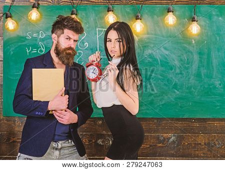 School Rules Concept. Man With Beard Hold Book And Sexy Girl Teacher Holds Alarm Clock, Chalkboard O