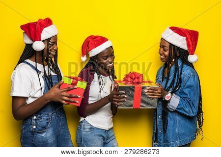 Three African Woman In Santa Hat Share Christmas Present On Yellow Background