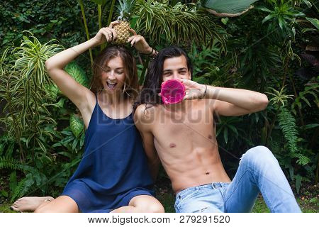 Beautiful Young Laughing Couple Are Sitting On The Grass With Tropical Fruits. Funny, Joke