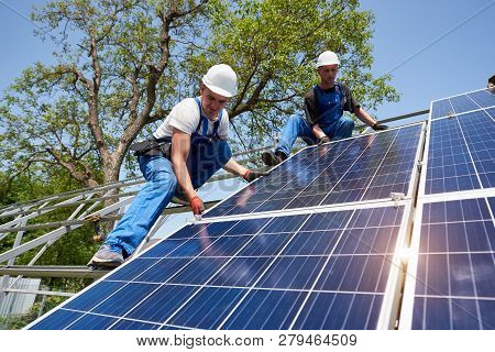 Two Young Technicians Installing Heavy Solar Photo Voltaic Panel On Tall Steel Platform On Green Tre