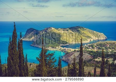 View To Assos Village In Sun Light And Beautiful Blue Sea. Cypress Trees In Foreground. Kefalonia Is