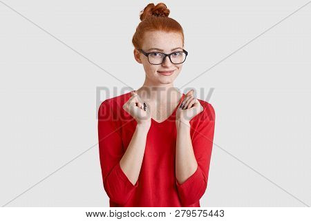 Happy Red Haired Freckled Young Woman With Knot Keeps Hands Pressed, Waits For Something With Intrig