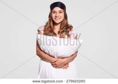 Indoor Shot Of Pleasant Looking Dark Haired Woman With Cheerful Expression Carries Pillow, Wears Eye