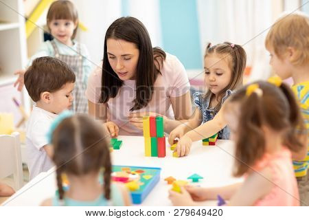 Kids Playing With Educational Toys At Table In Kindergarten. Nursery Teacher Looking After Children