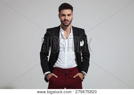 portrait of relaxed fashion man wearing black suit and red pants holding pockets while standing on light grey background