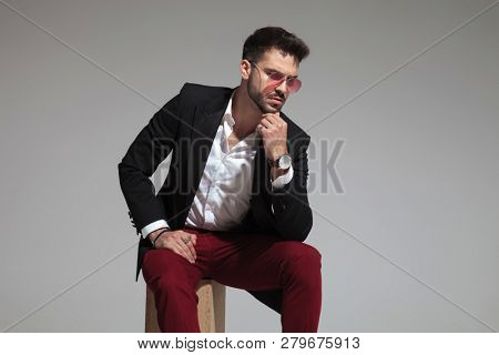pensive smart casual man sitting wearing red sunglasses and black suit looks down to side on light grey background
