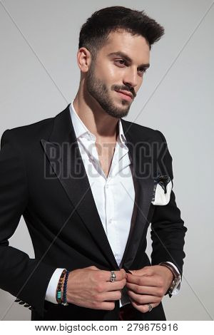 smiling handsome man buttoning black suit looks to side while standing on light grey background, portrait picture