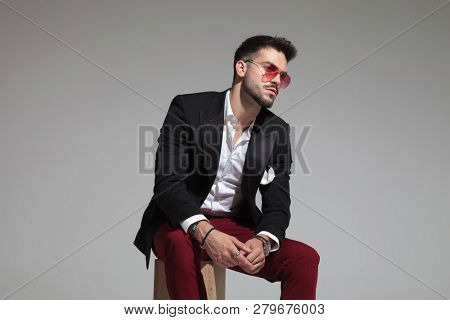 curious elegant man wearing black suit and burgundy pants looks to side while sitting on woden box, portrait picture