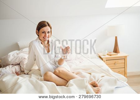 A Young Woman With Night Shirt Sitting Indoors On Bed In The Morning, Holding Water.