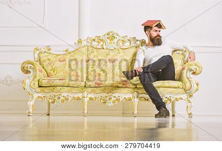 Macho Sits With Open Book On Head, Like Roof. Man With Beard And Mustache Sits On Sofa, White Wall B