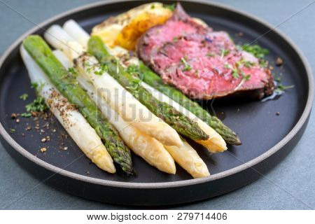 Traditional white and green asparagus with barbecue dry aged sliced beef fillet and fried potatoes served as closeup on a modern design plate 