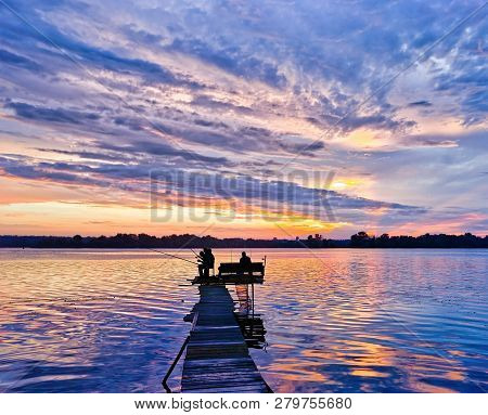 A Group Of Fishermen Fishing From The Bridge Against The Sunset Sky In The Clouds.