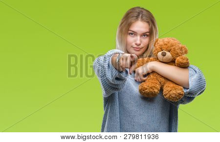 Young caucasian woman holding cute teddy bear over isolated background pointing with finger to the camera and to you, hand sign, positive and confident gesture from the front