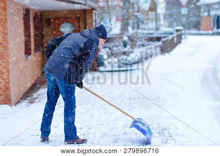 Man With Snow Shovel Cleans Sidewalks In Winter During Snowfall. Winter Time In Europe. Young Man In