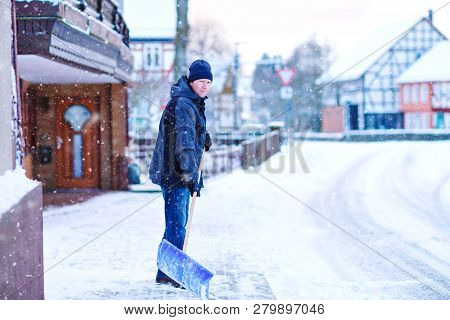 Man With Snow Shovel Cleans Sidewalks In Winter During Snowfall. Winter Time In Europe. Young Man In