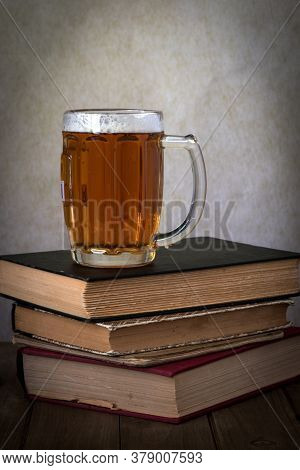 A Glass Of Golden Beer Over A Stack Of Hardbound Books On A Wooden Table Against Grey Background, Si