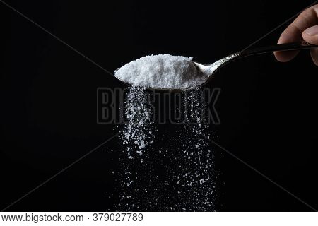 Salt Is Poured From A Spoon On A Black Background. Excessive Salt Intake. Coarse Salt