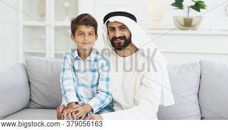 Portrait Of Young Handsome Muslim Father In Kandura Sitting On Couch In Living Room With Cute Small 