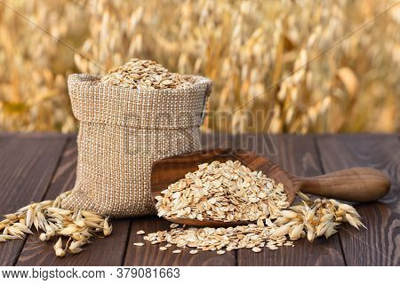 Raw Oatmeal In Bag And Wooden Scoop On Table With Ripe Cereal Field On The Background. Uncooked Porr