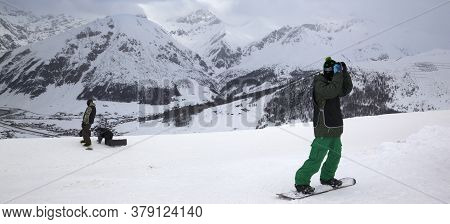 Snowboarders Starts On Off-piste Descent. High Snowy Mountains And Cloudy Storm Sky At Winter Evenin