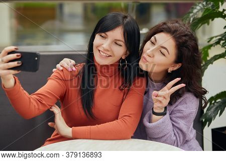 Two Young Women Taking Selfie In Cafe While Sitting At Table, Looking At Mobile Phone Camera, Smilin