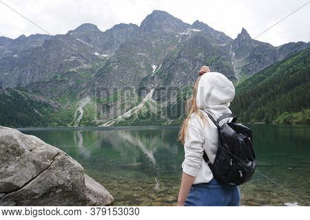A Girl In A White Jacket With A Black Backpack On Her Shoulders Stands Near A Lake In The Mountains