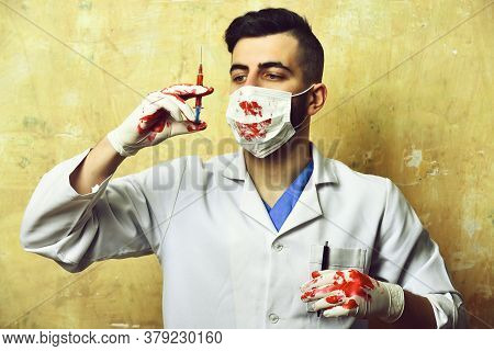 Doctor With Bloody Hands In Surgial Mask Looks At Syringe