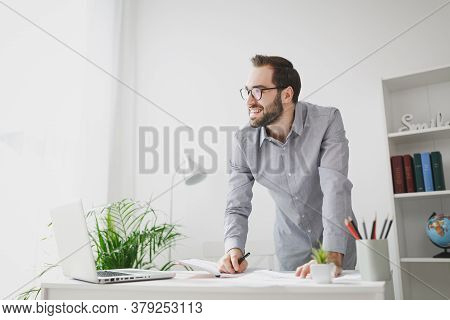 Smiling Young Bearded Business Man In Gray Shirt Standing Near Desk Working With Laptop Computer Pap