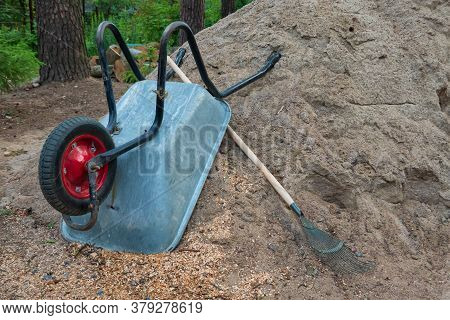 A Metal One-wheeled Wheelbarrow Lies Upside Down On A Pile Of Sand. There Are A Number Of Rake. Tree