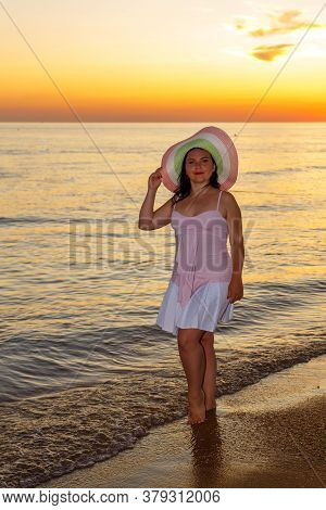 A Jewish Woman In A Hat And White Dress At Sunset Walks Along The Seashore.