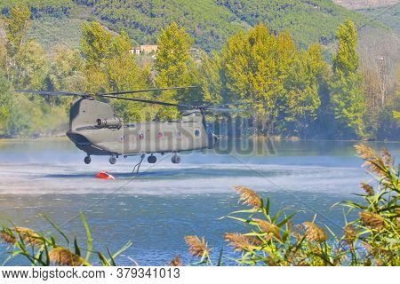 Fire Fighting Helicopter Of The Italian Army Flying Over A Lake To Collects Water In A Bucket To Ext