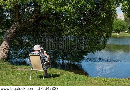 Elderly Woman In A White Hat Sits On A Folding Chair Near A Pond In A City Park And Solves A Crosswo
