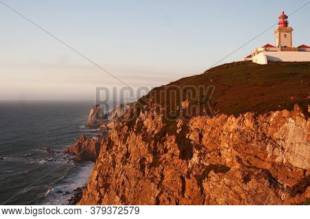 Lighthouse Standing On The Hige Rock In Front Of Ocean Coast In The Sunset Light.